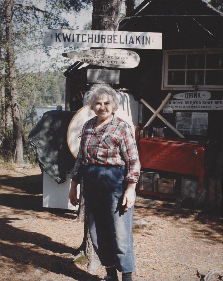 Dorothy Molter standing in front of her cabin with the "Kwitchurbeliakin" sign.