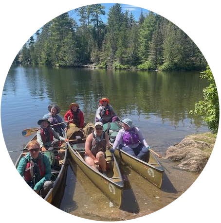 Three canoes floating on water near shore with seven women in them and a tree-lined shoreline behind them in the Boundary Waters